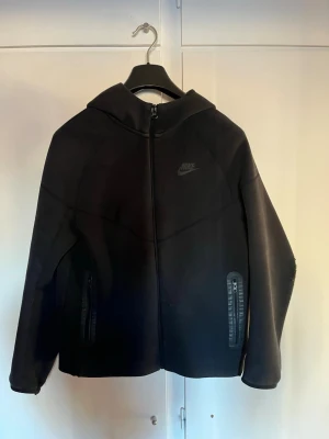 Svart Nike tech fleece  - Säljer en svart hoodie från Nike med hel dragkedja framtill och två fickor med dragkedja. Hoodien har en stilren design med Nike-logga på bröstet och en stor huva. Tillverkad i ett mjukt och stretchigt material som passar perfekt till streetwear. Storlek 158-170cm