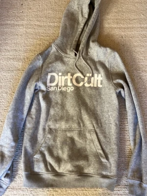 Grå hoodie Dirt Cult San Diego - Snygg grå hoodie från Dirt Cult med stor vit logga och texten 'San Diego' på bröstet. Klassisk känguruficka framtill, ribbade muddar och justerbar huva med dragsko. Perfekt för en avslappnad streetwear-look.