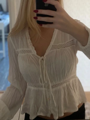 Vit boho-blus med knytning - Söt vit blus i boho-stil med volanger och knytning framtill. Blusen har v-ringning, spetsdetaljer och långärmade ärmar. Knappar och knytning framtill. Tillverkad i ett tunt och lätt tyg som ger en avslappnad känsla.