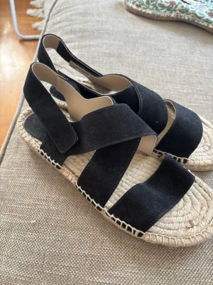 Svarta espadrillesandaler  - Helt NYa! Säljer ett par svarta sandaler med breda korslagda remmar i mockaimitation och öppen tå. Sulan är i klassisk flätad jute, vilket ger en avslappnad espadrillekänsla. Kardborreknäppning vid ankeln för enkel på- och avtagning. Perfekta för sommaren!