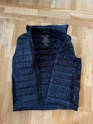 Svart dunväst från Calvin Klein - Snygg svart dunväst från Calvin Klein med hög krage och diskret logga längs sidan. Västen har två dragkedjeförsedda fickor och quiltad design. Perfekt att slänga över en hoodie eller tröja för extra värme. Materialet är lätt och smidigt, vilket gör den enkel att bära.