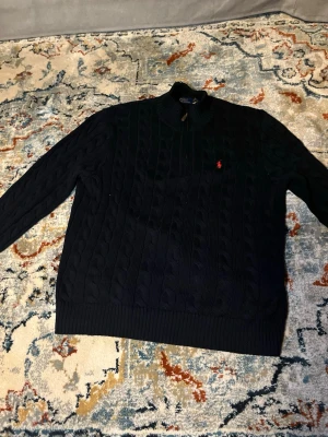 Mörkblå kabelstickad tröja Polo Ralph Lauren - Mörkblå kabelstickad tröja från Polo Ralph Lauren med halv dragkedja och klassisk röd logga på bröstet. Tröjan har ribbade muddar och hög krage, perfekt för lager på lager. Materialet ser ut att vara mjukt och värmande. Köpte den för 2100kr men säljer för 1300