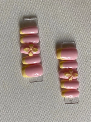 Press-on naglar rosa med gula tippar - Säljer ett set press-on naglar i ljusrosa med gula franska tippar och 3D-blommor som dekoration. Naglarna har en oval form och är gjorda i plast. Perfekt för dig som vill ha en söt och färgglad look med extra detaljer.