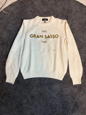 Vit stickad tröja Gran Sasso - Säljer en vit stickad tröja från Gran Sasso med rund halsringning och ribbade muddar. Tröjan har tryckt text '1952 Gran Sasso Italy' i guld på bröstet. Klassisk och enkel design som passar till mycket. Materialet känns mjukt och skönt.