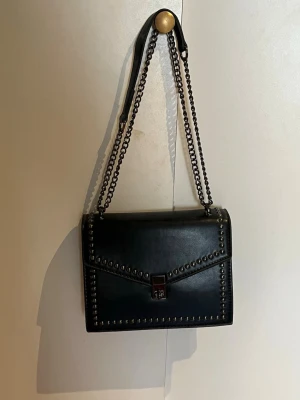 Svart axelväska med kedjerem - Snygg svart axelväska i läderimitation med coola nitar längs kanterna och en kedjerem i metall. Väskan har ett fyrkantigt format och stängs med ett metallspänne framtill. Perfekt för dig som gillar en edgy och stilren look.