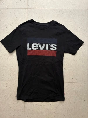 Svart Levi's t-shirt med logga - Säljer en svart t-shirt från Levi's i bomull med klassisk logga framtill i vitt, blått och rött. T-shirten har rund halsringning och korta ärmar. Perfekt för dig som gillar en enkel men snygg stil.