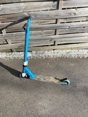Blå trick sparkcykel - Säljer en blå trick sparkcykel med svart handtag och mönstrad platta i beige och grått. Styret är i metall och hjulen är små och tåliga, perfekta för skateparken eller att cruisa runt på gatan.