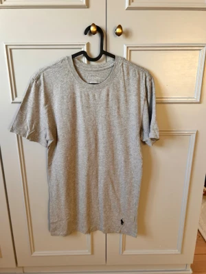 Grå t-shirt från Polo Ralph Lauren - Klassisk grå t-shirt från Polo Ralph Lauren med rund halsringning och korta ärmar. Tillverkad i mjuk bomull och har den ikoniska svarta polospelaren broderad nertill på framsidan. Perfekt basplagg för en clean och enkel stil.