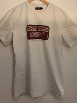 2st Vit Barbour International t-shirt - Säljer en vit t-shirt från Barbour International med tryck framtill i form av en brittisk flagga och logga. T-shirten har rund hals och korta ärmar. Tillverkad i mjuk bomull, perfekt för en avslappnad och stilren look.