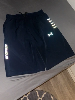 Svarta träningsshorts Under Armour - Snygga svarta träningsshorts från Under Armour med reflexdetaljer och logga på ena benet. Shortsen har snörning i midjan och är tillverkade i ett lätt och snabbtorkande syntetmaterial. Perfekta för gymmet eller löprundan.T-shirten är XL fast passar som L och shortsen är 2xl fast passar som xl 