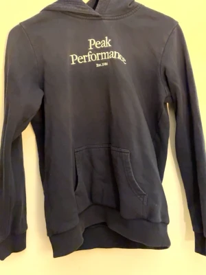 Svart hoodie från Peak Performance - Snygg svart hoodie från Peak Performance med vit logga och text på bröstet. Klassisk känguruficka framtill och ribbade muddar vid ärmar och nederkant. Tillverkad i mjukt bomullsmaterial och har en skön huva.