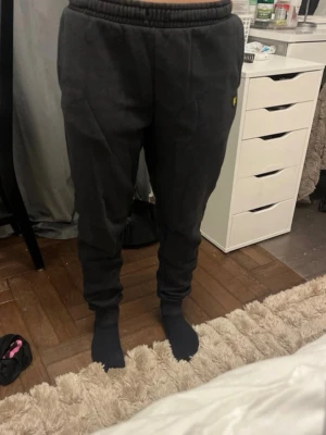 Svarta mjukisbyxor med resårmidja - Mörkgrå mjukisbyxor med resår i midjan och muddar vid bensluten. Byxorna har en bakficka och en liten gul logga på sidan. Tillverkade i mjukt och bekvämt material, perfekta för chill dagar hemma.