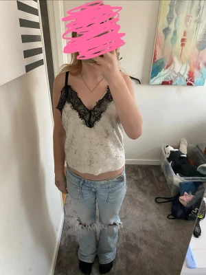 Ljusgrå linnetopp med spetsdetaljer - Säljer en ljusgrå linnetopp med tunna axelband och svart spets längs v-ringningen. Toppen har en croppad passform och är gjord i mjuk, glansig polyester som ger en lyxig känsla. Perfekt att styla med jeans eller kjol för en trendig look.