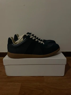 Mörkblå sneakers med gummisula - Snygga sneakers från Maison Margiela i en stilren mörkblå design med kontrasterande sula. Kombinationen av mocka och slätt läder ger en exklusiv och modern look. Perfekta både till vardag och mer uppklädda outfits.  Skorna är i fint skick och har en tidlös design som aldrig går ur stil. Bekväma att bära och enkla att matcha.    