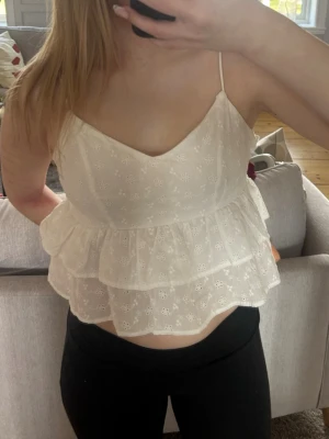Vit volang topp med broderade blommor - Söt vit linnetopp med tunna axelband och dubbla volanger. Toppen har små broderade blommor. Dragkedja i sidan. Knappt använd