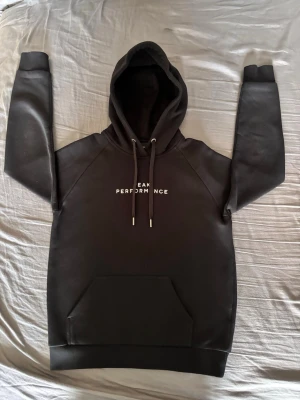 Svart Peak Performance hoodie - Snygg svart hoodie från Peak Performance med vit text på bröstet. Klassisk modell med stor huva, dragsnören och magficka framtill. Tillverkad i mjukt och tjockt material som känns riktigt skönt. Perfekt för dig som gillar stilrena plagg. Storlek M(i junior storlek), (inte vanlig M)