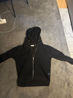 Svart zip hoodie från Lindbergh - Säljer en svart hoodie från Lindbergh med dragkedja framtill och två fickor. Klassisk modell med huva och långa ärmar. Perfekt till chill vardagsstil och enkel att matcha med det mesta. Materialet känns mjukt och bekvämt.