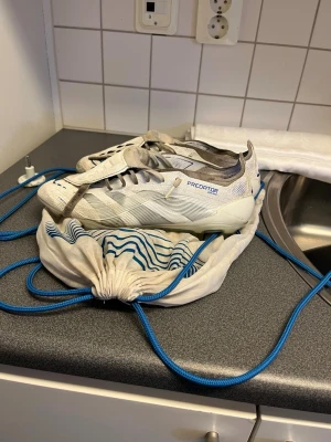 Adidas Predator vita fotbollsskor - Säljer ett par vita Adidas Predator elite FG fotbollsskor med grå och blå detaljer. Skorna har klassisk snörning och låg profil, tillverkade i syntetmaterial för bra grepp och kontroll på planen. Medföljer en vit tygpåse med blå snören och mönster.
