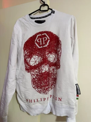 Vit Philipp Plein tröja med röd dödskalle - Cool vit långärmad tröja från Philipp Plein med stor röd dödskalle framtill och logga både fram och bak. Tröjan har ribbade muddar och rund hals. Perfekt för dig som gillar statement-plagg och streetwear vibes.