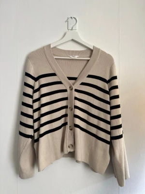 Beige randig kofta från Object - Beige kofta från Object med svarta horisontella ränder. Koftan har v-ringning, knäppning framtill med fyra knappar och ribbade muddar. Avslappnad passform och mjukt stickat material som känns skönt mot huden.