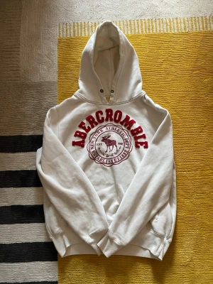 Vit hoodie från Abercrombie med tryck - Vit hoodie från Abercrombie med rött broderat tryck och logga på bröstet. Klassisk känguruficka framtill och huva med dragsko. Mjuk insida och ribbade muddar vid ärmar och nederkant. Perfekt för en chill och sportig look.