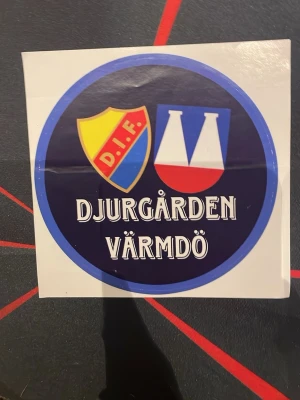 Djurgården Värmdö klistermärke sofialäktaren  - Klistermärke med Djurgården Värmdö-tryck. Rund design med blå bakgrund, DIF-sköld i gult, rött och blått samt två vita och röda symboler. Texten 'DJURGÅRDEN VÄRMDÖ' i vitt. Perfekt för att visa ditt stöd.
