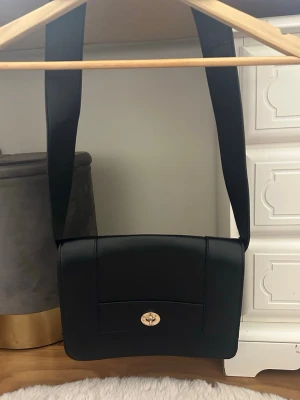 Svart axelväska med bred rem - Stilren svart axelväska i slätt konstläder med bred axelrem och guldfärgat vridlås framtill. Väskan har en fyrkantig form och minimalistisk design som passar till många olika outfits. Perfekt för dig som gillar enkel men snygg stil.