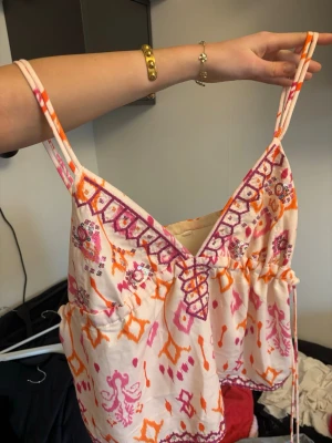 Mönstrad linnetopp med broderi - Säljer en färgglad linnetopp med tunna axelband och v-ringning. Toppen har ett rosa, orange och vitt mönster samt broderade detaljer längs ringningen. Perfekt för varma dagar och ger en bohemisk vibe.