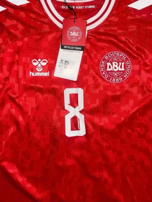 Röd Danmark fotbollströja Hummel - Snygg röd Danmark fotbollströja från Hummel med vita detaljer och nummer 8 på bröstet. Tröjan har DBU-logga, rund hals med vita ränder och tryckta märken på ärmarna. Tillverkad i återvunna fibrer och lätt funktionsmaterial som andas and I take PayPal