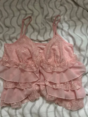 Rosa spetsbody med volanger - Söt rosa body med tunna axelband, spetsdetaljer och flera lager volanger framtill. Materialet är en kombination av spets och mjuk textil som ger en romantisk och feminin känsla. Perfekt för dig som gillar gulliga och lekfulla underkläder.