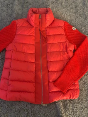 Röd pufferjacka från Moncler - Snygg röd pufferjacka från Moncler med stickade ärmar och hög krage. Jackan har quiltad kropp, dragkedja framtill och Moncler-logga på ena ärmen. Perfekt för kalla vinterdagar och ger en sportig look.
