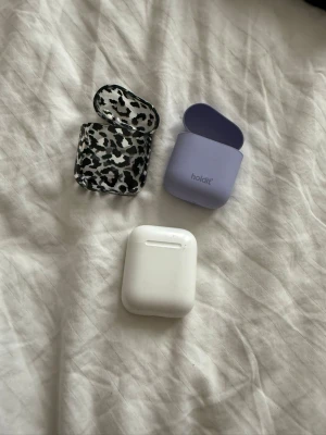 Apple AirPods med fodral och skal - Säljer ett par Apple AirPods 2. Medföljer två extra skal: ett lila silikonfodral från Holdit och ett svartvitt leopardmönstrat skal. Perfekt för dig som vill ha snygga och skyddade hörlurar i fickan.