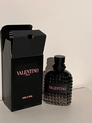 Valentino Uomo Born In Roma parfym - Valentino Uomo Born In Roma parfym i en snygg svart flaska med nitar och rosa text. Flaskan har en unik, lyxig design med mattsvart lock och detaljer. Kommer i matchande svart kartong med rosa logga. Perfekt för dig som gillar exklusiva dofter och cool design.