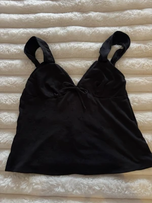 Svart linnetopp med knyt fram och bak  - Svart linnetopp med breda axelband och v-ringning. Toppen har en söt knyt-detalj framtill och diskret broderat blommönster i tyget. Perfekt att styla med jeans eller kjol för en trendig look.