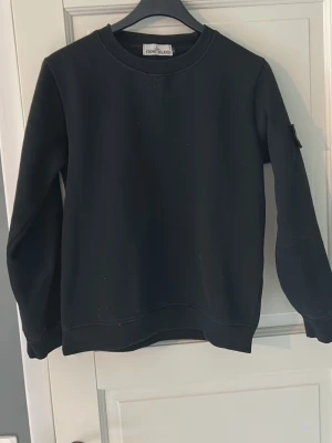 Svart sweatshirt från Stone Island - Säljer en svart sweatshirt från Stone Island med klassisk rund hals och ribbade muddar. Tröjan har den ikoniska Stone Island-loggan på vänster ärm. Tillverkad i mjuk bomull för skön komfort och stilren look.