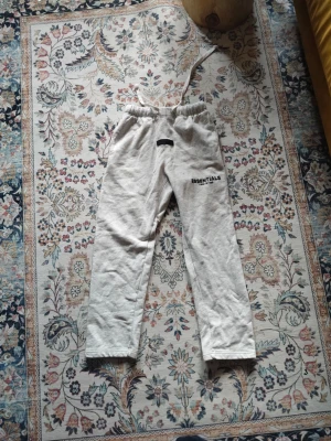 Grå Essentials mjukisbyxor Fear of God - Säljer ett par ljusgrå mjukisbyxor från Fear of God Essentials. Byxorna har rak passform, resår i midjan med dragsko och svart Essentials-logga tryckt på vänster lår. Tillverkade i mjukt bomullsmaterial för maximal komfort.