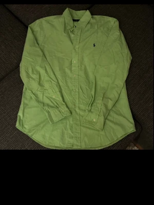 Ralph Lauren Oxford Skjorta - Snygg grön skjorta från Ralph Lauren med klassisk krage och blå broderad logga på bröstet. Skjortan har knappar framtill och långa ärmar. Perfekt för dig som vill ha en stilren och tidlös look.