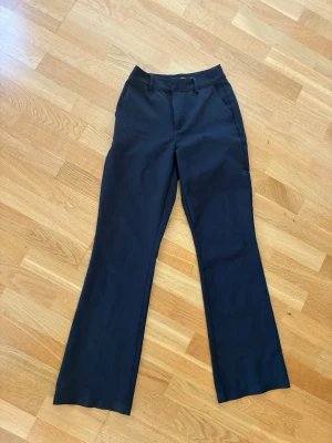 Svarta utsvängda kostymbyxor - Snygga svarta kostymbyxor med utsvängda ben. Klassisk design med bälteshällor och sidofickor. Perfekta för en stilren och trendig look. Materialet känns mjukt och följsamt, vilket ger en bekväm passform.