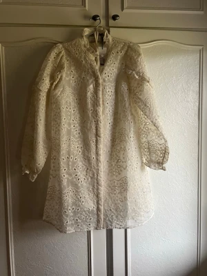 Vit broderad klänning från Neo Noir - Broderad kortklänning från Neo Noir i ivory-vit nyans. Klänningen har långärmade ärmar, hög krage och vackra spetsdetaljer över hela plagget. Perfekt för dig som gillar romantiska och detaljerade klänningar med elegant känsla.