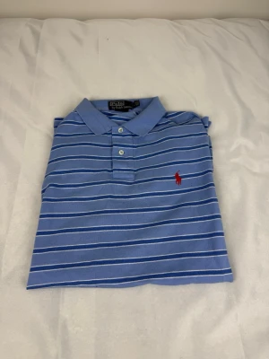 Blå randig piké från Ralph Lauren - Klassisk blå pikétröja från Ralph Lauren med vita ränder och röd broderad logga på bröstet. Tröjan har krage, knappar framtill och är tillverkad i mjuk bomull. Perfekt för en avslappnad men stilren look.