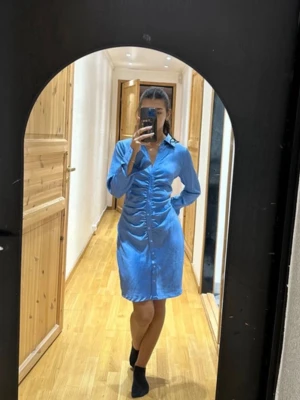 Blå omlottklänning från New Look - Snygg blå kortklänning från Neo Noir i glansigt material med lång ärm och v-ringning. Klänningen har knappar framtill, markerad midja med rynkade detaljer och klassisk krage. 