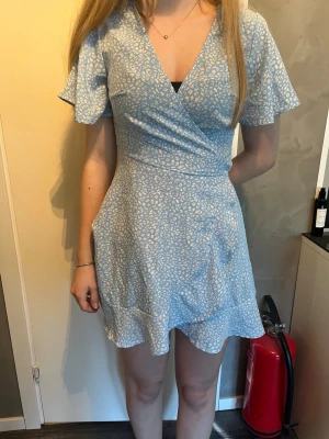 Blå omlottklänning med mönster - Söt blå kortklänning med vitt mönster och omlottdesign. Klänningen har korta ärmar, v-ringad hals och volangkant nertill. Perfekt för varma dagar och ger en fräsch look. Materialet känns lätt och luftigt. Säljs tillsammans med en grön likadan