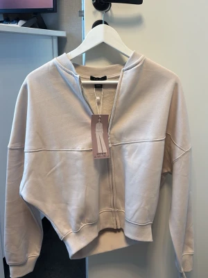 Beige zip hoodie från A.M.S.U - Stilren beige hoodie med dragkedja från A.M.S.U. Hoodien har en croppad passform, ribbade muddar och mjukt material. Perfekt för en avslappnad och trendig look. Enkel design utan tryck eller mönster. Aldrig använt, prislapp med. Bomberjacka vibe