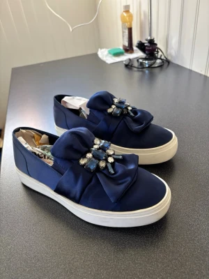 Mörkblå slip-on sneakers med rosett - Snygga mörkblå slip-on sneakers med stor satängrosett och glittrande stenar på ovansidan. Skorna har en tjock vit platåsula och är tillverkade i ett glansigt tygmaterial. Perfekta för dig som vill sticka ut med en unik detalj på fötterna.