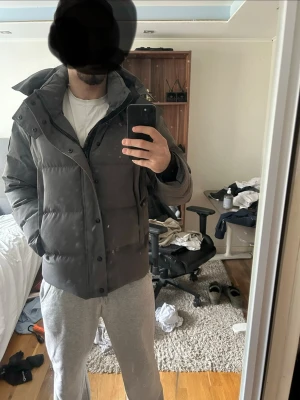 Mörkgrå dunjacka från Canada Goose - Säljer en mörkgrå dunjacka från Canada Goose med hög krage och avtagbar huva. Jackan har en patch med logga på ärmen, stora framfickor och stängs med både dragkedja och knappar. Perfekt för kalla vinterdagar och riktigt varm.