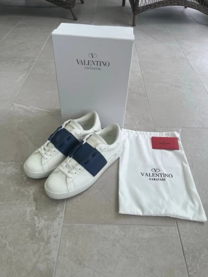 Valentino Garavani vita sneakers - Snygga vita sneakers från Valentino Garavani med breda mörkblå band över vristen och vita skosnören. Skorna har en stilren och modern design med guldfärgad logga på plösen. Tillverkade i läder och levereras med originalkartong och dustbag.
