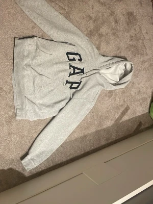 Ljusgrå hoodie från GAP - Ljusgrå hoodie från GAP med stor logga i svart och vitt på bröstet. Klassisk modell med huva och känguruficka framtill. Tillverkad i mjukt sweatshirtmaterial, perfekt för en avslappnad stil.