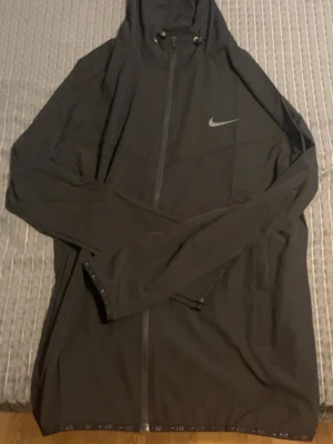 Svart Nike Pro vindjacka med huva - Snygg svart vindjacka från Nike Pro med hel dragkedja, huva med dragsko och två sidofickor. Jackan har diskret Nike-logga på bröstet och 'NIKE PRO' tryckt på ryggen. Tillverkad i lätt och vindtätt material, perfekt för aktiva dagar utomhus.