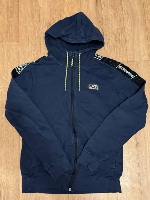 Marinblå hoodie från EA7 Emporio Armani - Snygg marinblå hoodie med dragkedja från EA7 Emporio Armani. Har huva med gula snören och logga på bröstet samt detaljer på axlarna med EA7- och Armani-tryck. Tillverkad i mjukt bomullsmaterial, perfekt för en avslappnad och trendig look.
