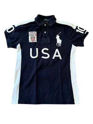 Ralph Lauren USA pikétröja - Snygg mörkblå pikétröja från Ralph Lauren med vita detaljer och stora USA-tryck fram och bak. Äkta såklart!
